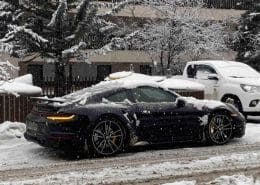 Black Porsche 911 Turbo in snowy residential street with white pickup truck nearby.